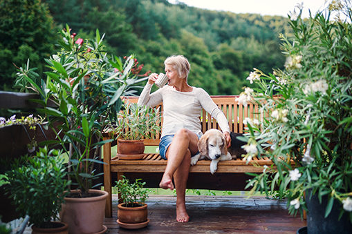 Woman Sitting on Bench with Dog, Drinking Coffee Surrounded by Plants