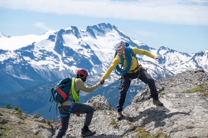 One Hiker Holding the Hand of Another and Helping them Up a Mountain