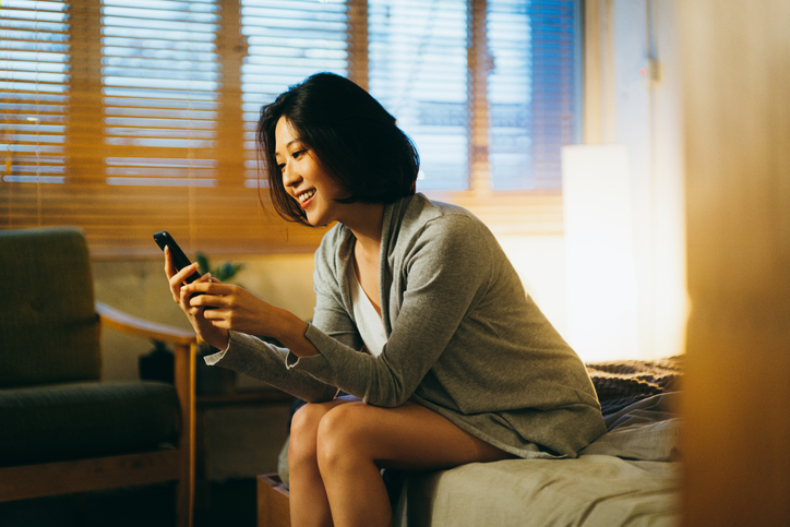 Woman Smiling and Sitting on Side of Bed Using Cell Phone